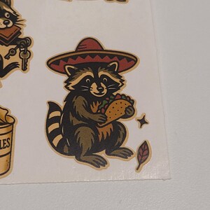 May include: A sheet of stickers featuring cartoon raccoons. One raccoon wears a sombrero and holds a taco. Other raccoons are depicted with keys, in a trash can, and with a tail. The stickers have a vintage, hand-drawn style.