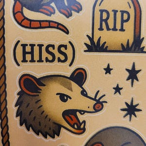 May include: A sheet of tattoo-style stickers featuring a possum with an open mouth, the word "HISS", a tombstone with "RIP", and stars. The possum is brown with red accents. The stickers have a vintage aesthetic.