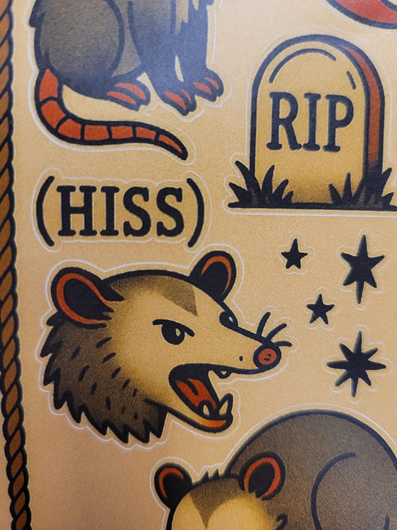 May include: A sheet of tattoo-style stickers featuring a possum with an open mouth, the word "HISS", a tombstone with "RIP", and stars. The possum is brown with red accents. The stickers have a vintage aesthetic.