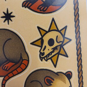 May include: Sheet of stickers featuring opossums, a skull, and starburst designs. The opossums are brown with orange feet and tails. The skull is yellow with a black eye socket. The background is tan.