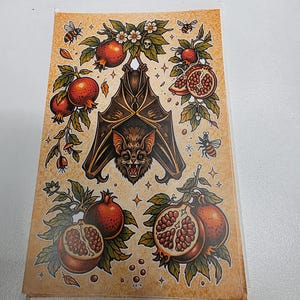May include: A decorative sticker featuring a bat with outstretched wings, surrounded by pomegranates, bees, and floral elements. The design has a vintage, tattoo-inspired aesthetic with a brown and red color palette.