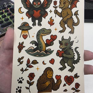 May include: A sheet of temporary tattoos featuring various designs. The designs include a moth with a heart, a devil holding a rose, a UFO, a Loch Ness Monster, a green demon, a Bigfoot, roses, hearts, and paw prints.