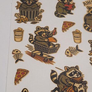 May include: A sheet of stickers featuring cartoon raccoons in various poses. One raccoon is in a trash can, another holds a heart, and others are eating pizza and noodles. The stickers have a vintage, hand-drawn style with brown, tan, and olive green colors.