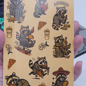 May include: A sheet of raccoon stickers in various poses, holding pizza, beer, and noodles. The raccoons are depicted in a cartoon style, with brown and gray fur and black stripes. Some stickers have the text "AHHHHH" and "NOODLES".