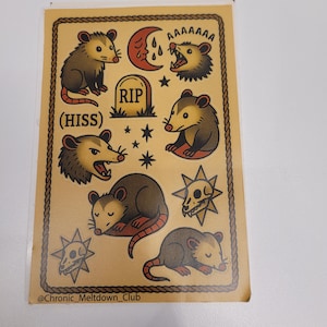 May include: A sticker sheet featuring opossum illustrations in a vintage tattoo style. The opossums are depicted in various poses, with text that reads "AAAAAAA" and "(HISS)". The design includes a RIP tombstone, stars, and a crescent moon.