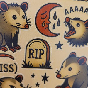 May include: A sheet of stickers featuring opossum illustrations in a traditional tattoo style. Designs include opossums, a crying moon, a tombstone with "RIP", and stars. The color palette is primarily tan, brown, and red.