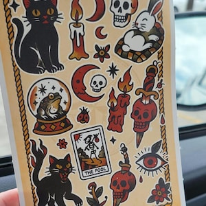 May include: A sticker sheet with various tattoo-style illustrations. Designs include a black cat, skull, rabbit, candles, a snow globe with a frog, a tarot card, and other mystical symbols. The sheet is bordered with a rope design.