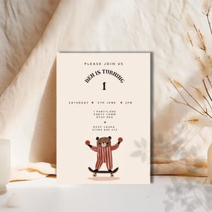 Könnte beinhalten: Eine beige Einladungskarte mit braunem Text, der lautet: "Bitte schließen Sie sich uns an Ben wird 1 Samstag, 5. Juni 14:00 Uhr 1 Partylane Party Town KY29 8GH RSVP Laura 07564 828 372". Ein brauner Cartoon-Bär, der einen rot-weiß gestreiften Pyjama trägt, fährt Skateboard mit roten Bällen in seinen Händen.