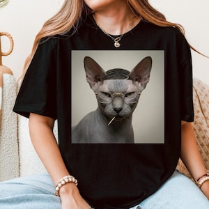 Pode incluir: Camiseta preta com um gráfico de um gato Sphynx usando óculos e um palito de dentes. O gato tem a cabeça raspada e uma expressão séria. O gato está centralizado na camiseta.
