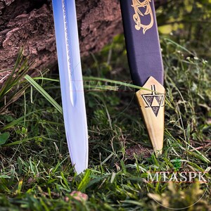 Puede incluir: Primer plano de la punta de una espada plateada con una empu&ntilde;adura dorada. La empu&ntilde;adura presenta un dise&ntilde;o triangular con un s&iacute;mbolo en su interior. La espada descansa sobre hierba verde.