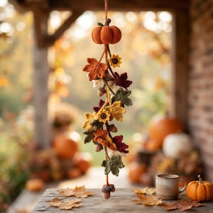 May include: A hanging crochet decoration featuring a central orange pumpkin at the top, followed by a vertical arrangement of crocheted autumn leaves in shades of red, orange, yellow, and green, small sunflowers, and brown acorns on a string.