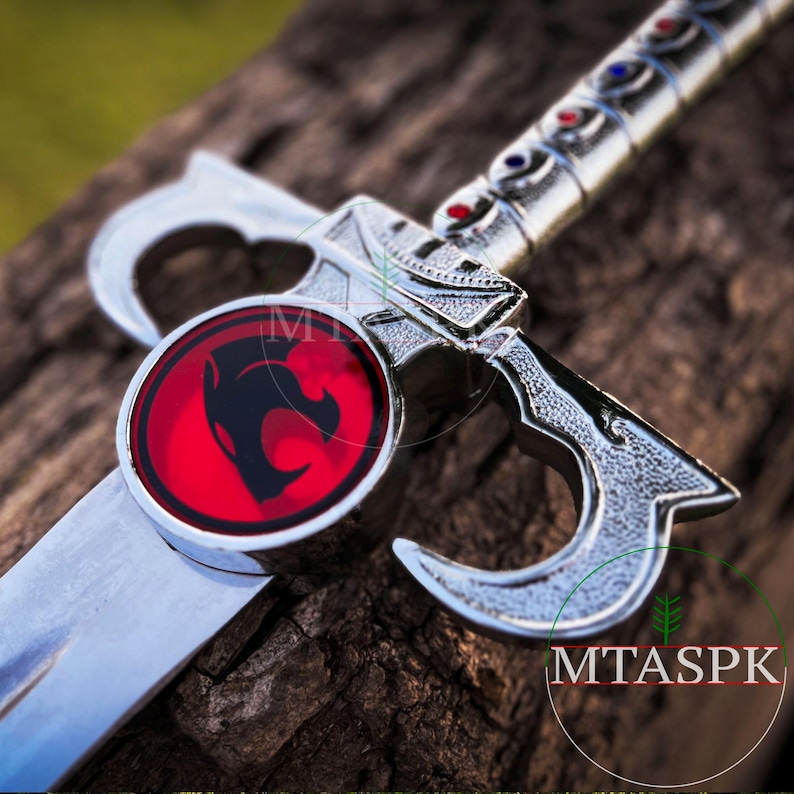 May include: Close-up of a silver sword with a red and black emblem featuring a stylized cat head. The emblem is surrounded by a silver ring.
