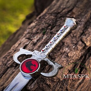 May include: A silver toy sword with a red and silver lion emblem on the hilt. The sword has a silver blade and a silver handle with red and blue accents.