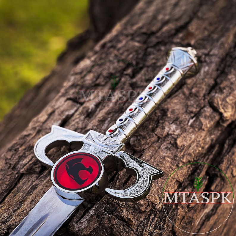 May include: A silver toy sword with a red and silver lion emblem on the hilt. The sword has a silver blade and a silver handle with red and blue accents.