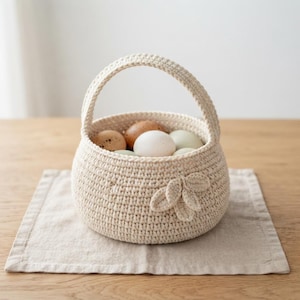 May include: A round, cream-coloured crocheted basket with a single arched handle. The exterior features a decorative cluster of three crocheted leaves on the side. The basket is filled with a collection of natural, speckled, and solid-coloured eggs.