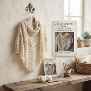 Può includere: Uno scialle all'uncinetto color crema con frange appeso a un gancio da parete decorativo. Accanto ci sono due libretti di modelli intitolati Aurora Borealis Shawl, un gomitolo di lana, un uncinetto in legno e una tazza contenente forbici.