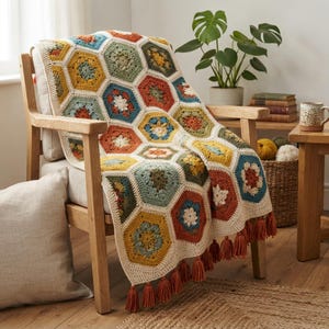 Può includere: Una coperta all'uncinetto con un motivo di esagoni multicolori nelle tonalità giallo senape, verde salvia, terracotta e blu, uniti da un filo color crema. La coperta è rifinita con nappe coordinate color crema lungo il bordo.