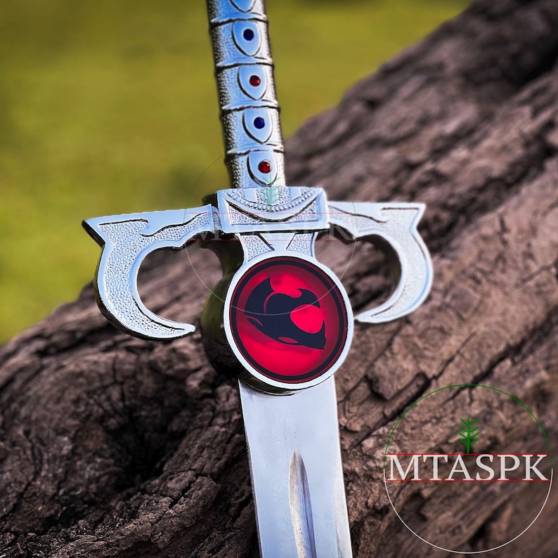 May include: A silver sword with a red and black emblem on the pommel. The emblem features a stylized animal with a red background. The sword is resting on a piece of wood.
