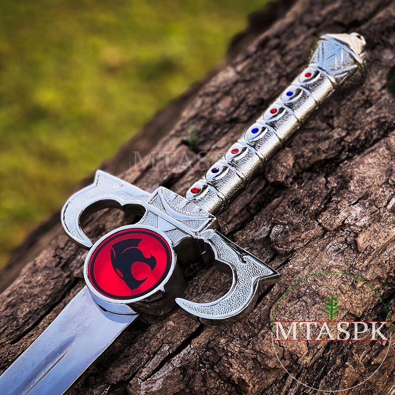 May include: A silver sword with a red and black lion emblem on the pommel. The sword has a silver handle with blue and red accents.