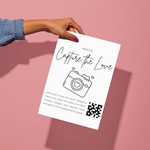 May include: A white card with black text that says "Help us Capture the Love". There is a black and white illustration of a camera with a heart inside. Below the camera is a QR code.