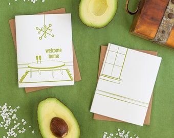 May include: Two blank greeting cards with brown envelopes. The card on the left has a green illustration of a table with two glasses and a chandelier. The text on the card reads "welcome home". The card on the right is blank. The cards are on a green surface with two avocado halves and white pebbles.