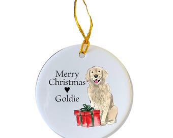 Puede incluir: Adorno navideño de cerámica blanca con un perro golden retriever sentado junto a una caja de regalo roja. El adorno tiene el texto "Merry Christmas" y un corazón con el nombre "Goldie" escrito debajo.