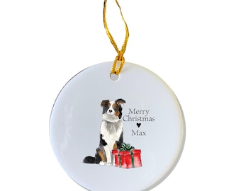 Puede incluir: Adorno navideño de cerámica blanca con un perro border collie blanco y negro sentado junto a una caja de regalo roja. El adorno tiene el texto "Feliz Navidad" y un símbolo de corazón, seguido del nombre "Max".