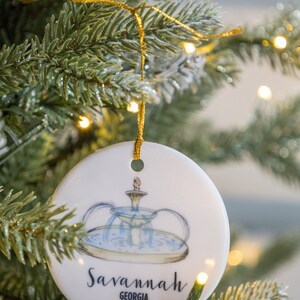 Puede incluir: Adorno navide&ntilde;o redondo blanco con una ilustraci&oacute;n azul y verde de una fuente y el texto "Savannah, Georgia".