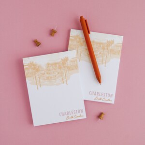 May include: Two white notepads with a sketch of a fountain and palm trees. The notepads have the text "Charleston, South Carolina" printed on them. A red pen and two wooden push pins are also in the image.