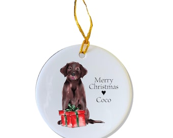 Puede incluir: Un adorno navideño de cerámica blanca con un labrador retriever marrón que sostiene una caja de regalo roja y verde. El adorno tiene el texto "Feliz Navidad" y "Coco".