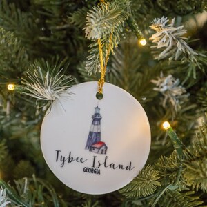 Puede incluir: Adorno navide&ntilde;o de cer&aacute;mica blanca con una ilustraci&oacute;n de un faro y el texto "Tybee Island, Georgia".