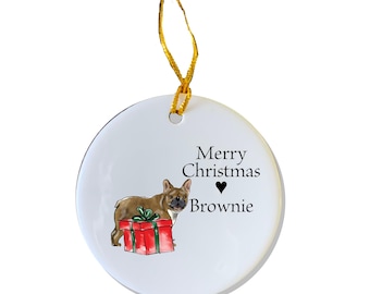 Puede incluir: Un adorno navideño de cerámica blanca con un bulldog francés marrón y blanco sentado en una caja de regalo roja. El adorno tiene el texto "Merry Christmas" y un símbolo de corazón, seguido del nombre "Brownie".