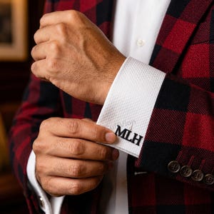 May include: A pair of black custom monogram cufflinks featuring the letters MLH in a bold serif font, attached to a white textured dress shirt cuff, worn under a red and black buffalo plaid patterned blazer with dark buttons.