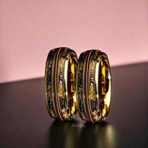 May include: Two gold-colored rings with intricate detailing. Each ring features alternating bands of gold, black, and a textured gold-colored material. The rings are displayed against a pink and black background. The text "Sutter & Crow Jewels" is visible.