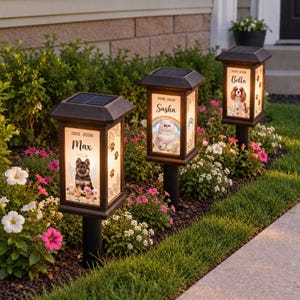 May include: Three black solar-powered garden lanterns featuring illuminated panels with pet portraits, names, and dates. Each lantern displays a different animal, including a German Shepherd, a cat, and a spaniel, surrounded by floral graphics.