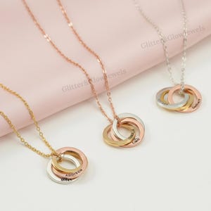 Peut inclure: Trois colliers personnalisés avec des anneaux entrelacés en or, or rose et argent. Chaque collier présente des noms gravés sur les anneaux. Les colliers sont présentés sur une surface rose, mettant en valeur le design.