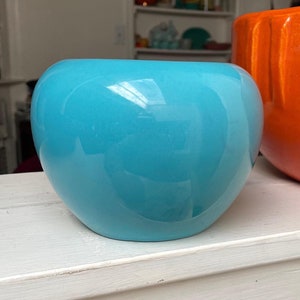 May include: Two ceramic planters, one in a bright orange and the other in a turquoise blue. The planters are both round and have a smooth, glossy finish.