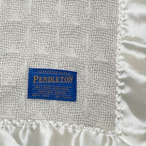 May include: An off-white wool blanket with a textured grid pattern and a smooth satin border. A rectangular blue label is stitched onto the fabric, featuring the text Pendleton, 100% Pure Virgin Wool, Portland, Oregon, Since 1863, Dry Clean.