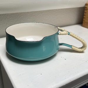 May include: A teal enamel bowl with a smooth, rounded shape and a cream-colored interior. The bowl features a single metal handle wrapped in light-colored woven rattan or wicker material, with a small notch along the top rim of the bowl.