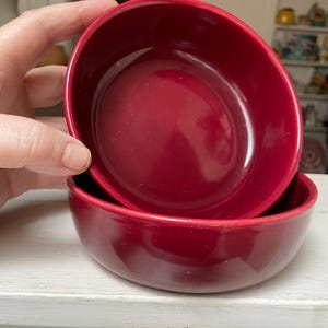 May include: Two red ceramic bowls, one stacked on top of the other. The bowls have a glossy finish and a rounded shape. The bowls are sitting on a white surface.