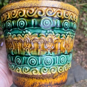 May include: A vintage ceramic planter with a textured, layered design. The planter features bands of green, yellow, and brown with repeating scroll and circle patterns. The rim and base have a speckled finish.