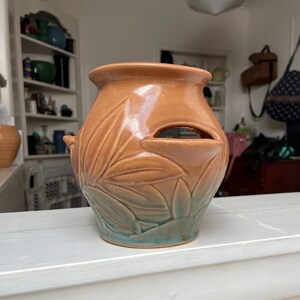 May include: A brown ceramic planter with a green glaze and a leaf design. The planter has a hole in the side for drainage.