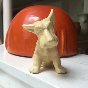 May include: A small, white ceramic figurine of a dog, possibly a Scottish Terrier, sitting on a white surface in front of a large, orange ceramic bowl.