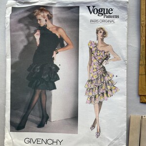 May include: A vintage Vogue Patterns sewing pattern featuring two illustrations of Givenchy dresses. One dress is black with a ruffled skirt, and the other is a floral print with a one-shoulder design. The pattern number is 1702.