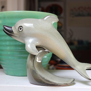 May include: A ceramic dolphin figurine in a light gray color with a dark gray tail. The dolphin is leaping out of the water, with its tail curled up behind it. The figurine is sitting on a table next to a green ceramic planter.