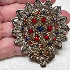 May include: An ornate, circular brooch with a sunburst design. The brooch features a central design with red and blue jewels, surrounded by gold and silver embroidery. The outer edge has silver-colored petal-shaped elements.