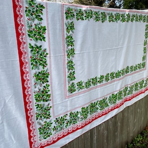 May include: White Christmas tablecloth with a red border and a holly and berry design. The border features a red lace trim. The holly and berry design is green with red berries. The tablecloth is hanging on a wooden fence.