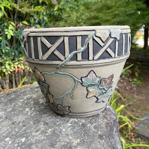 May include: A round ceramic planter with a matte gray finish. The upper rim features a black band with a raised lattice pattern. The body is decorated with a raised, vine-like relief design featuring green and brown leaves wrapping around the exterior.
