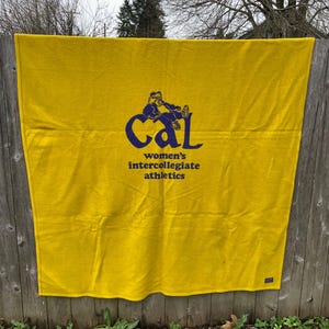 May include: A golden yellow blanket with the text "Cal women's intercollegiate athletics" in navy blue. The blanket is hanging on a wooden fence outdoors.