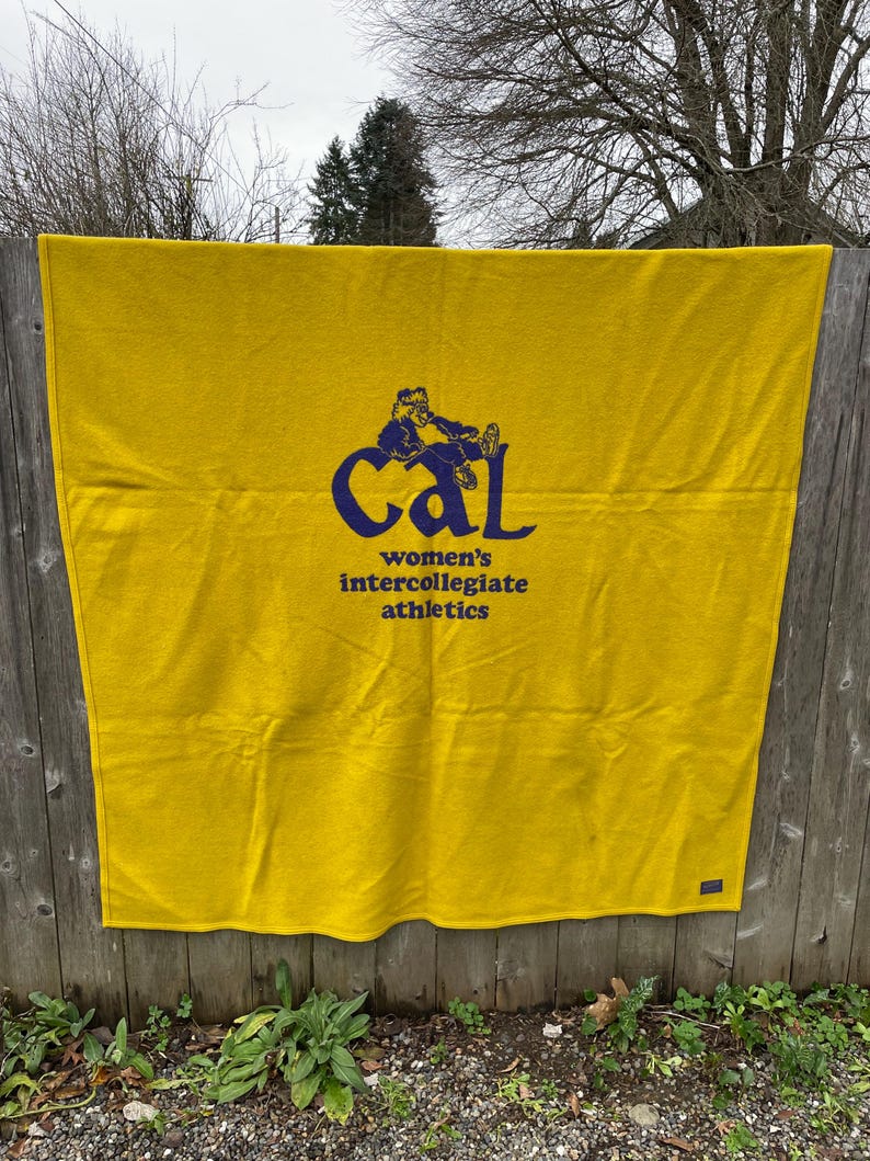 May include: A golden yellow blanket with the text "Cal women's intercollegiate athletics" in navy blue. The blanket is hanging on a wooden fence outdoors.